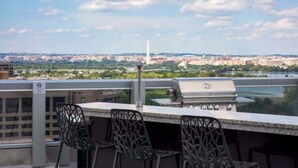 An outdoor bar area with a grill, chairs, and a view of a cityscape including a tall monument.