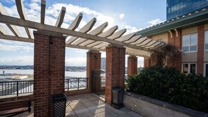 Terraço no telhado com pérgola, pilares de tijolo, vista para um corpo d'água e um edifício com janelas.