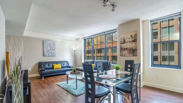 Uma sala de estar moderna com sofá de couro preto, mesa de jantar de vidro com cadeiras e grandes janelas com vista para um edifício.