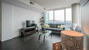A modern living room with a flat-screen TV, a glass coffee table, a sofa with cushions, and a large window offering a city view. - Downtown 2br Apartment Near Convention Center 2 Bedroom Apts by RedAwning (Baltimore)