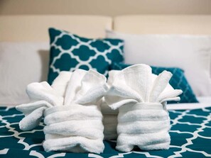 A bed with a teal and white patterned bedspread, two white towels arranged in a decorative manner, and a blue pillow with a white pattern.