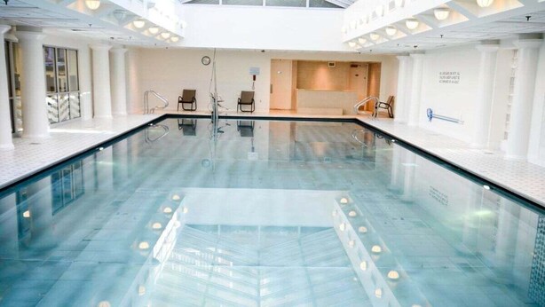 Ein großes Hallenbad mit klarem Wasser, umgeben von weißen Wänden und Säulen, ausgestattet mit Sitzgelegenheiten und einem Umkleideraum.