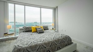 A bedroom with a large window, a bed with patterned bedding, and a view of a cityscape.
