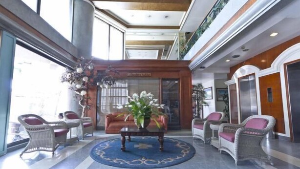 A lobby with wicker chairs, a central table with a vase of flowers, and a blue patterned rug.