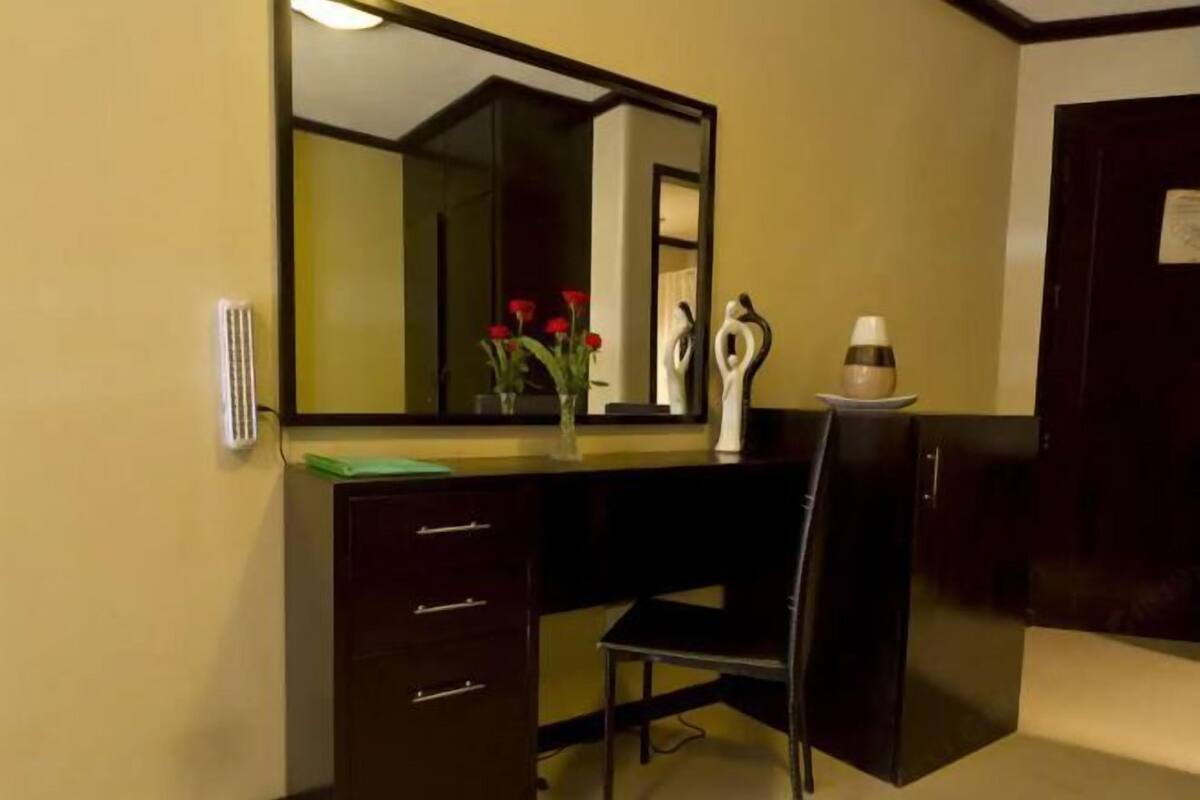 Um quarto com uma escrivaninha de madeira escura, uma cadeira, um espelho grande e um pequeno vaso com flores vermelhas.