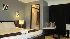 A hotel room with a large bed, a bedside table with a lamp, a small seating area with a chair and cushions, and a mirror on the wall.
