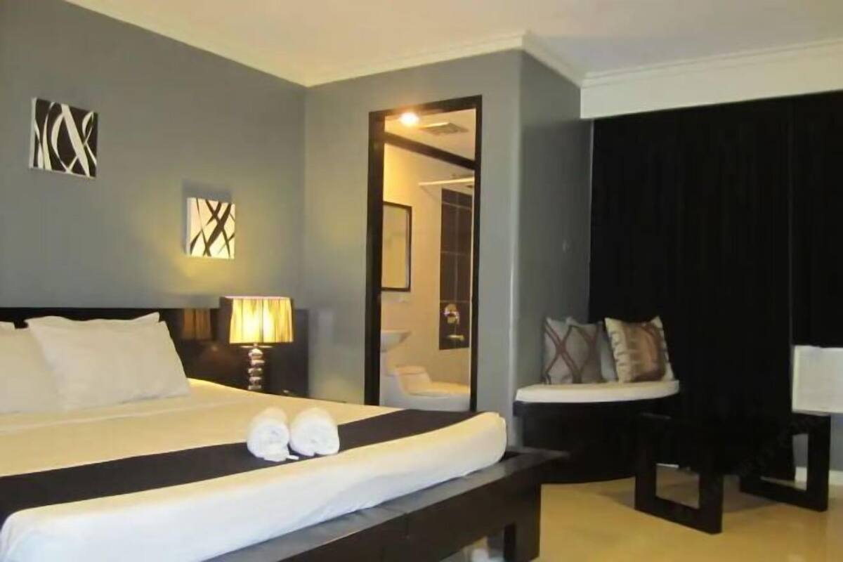Quarto de hotel com cama grande, mesa de cabeceira com abajur, um espaço de estar com poltrona e almofadas, e um espelho na parede.