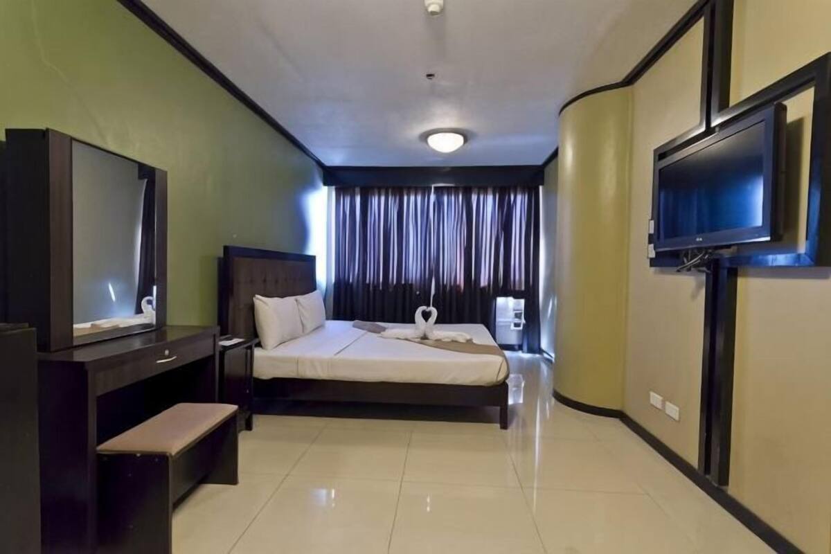 Quarto de hotel com cama, escrivaninha, televisão e janela com cortinas.