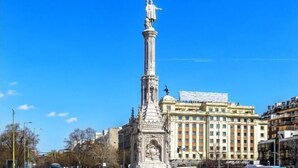 A tall, ornate monument with a statue at the top, surrounded by urban buildings and a clear blue sky.