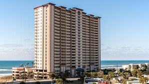 Un hotel alto de varios pisos con balcones, situado cerca de una playa con palmeras y cielo despejado.