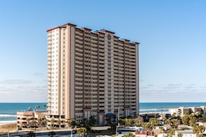Un hotel alto de varios pisos con balcones, situado cerca de una playa con palmeras y cielo despejado.