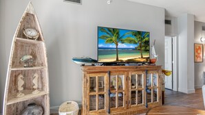 Una sala de estar con un mueble de madera, un televisor mostrando una escena de playa y un estante decorativo con un pez y un caballito de mar.