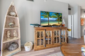 Una sala de estar con un mueble de madera, un televisor mostrando una escena de playa y un estante decorativo con un pez y un caballito de mar.