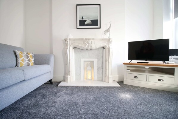 Ein Wohnzimmer mit grauem Sofa, einem weißen Kamin, einem Flachbildfernseher auf einem Holzboden und einem gerahmten Bild an der Wand.