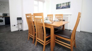 A dining area with a wooden table and chairs, a painting on the wall, and a kitchen in the background.
