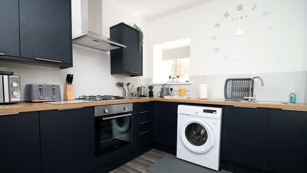 A modern kitchen with dark cabinetry, a built-in oven, a washing machine, and a clock on the wall.