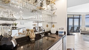 A kitchen with a granite countertop, a blender, and a glass wine rack with decorative items. - Phoenix West 1108 by Liquid Life (Orange Beach)