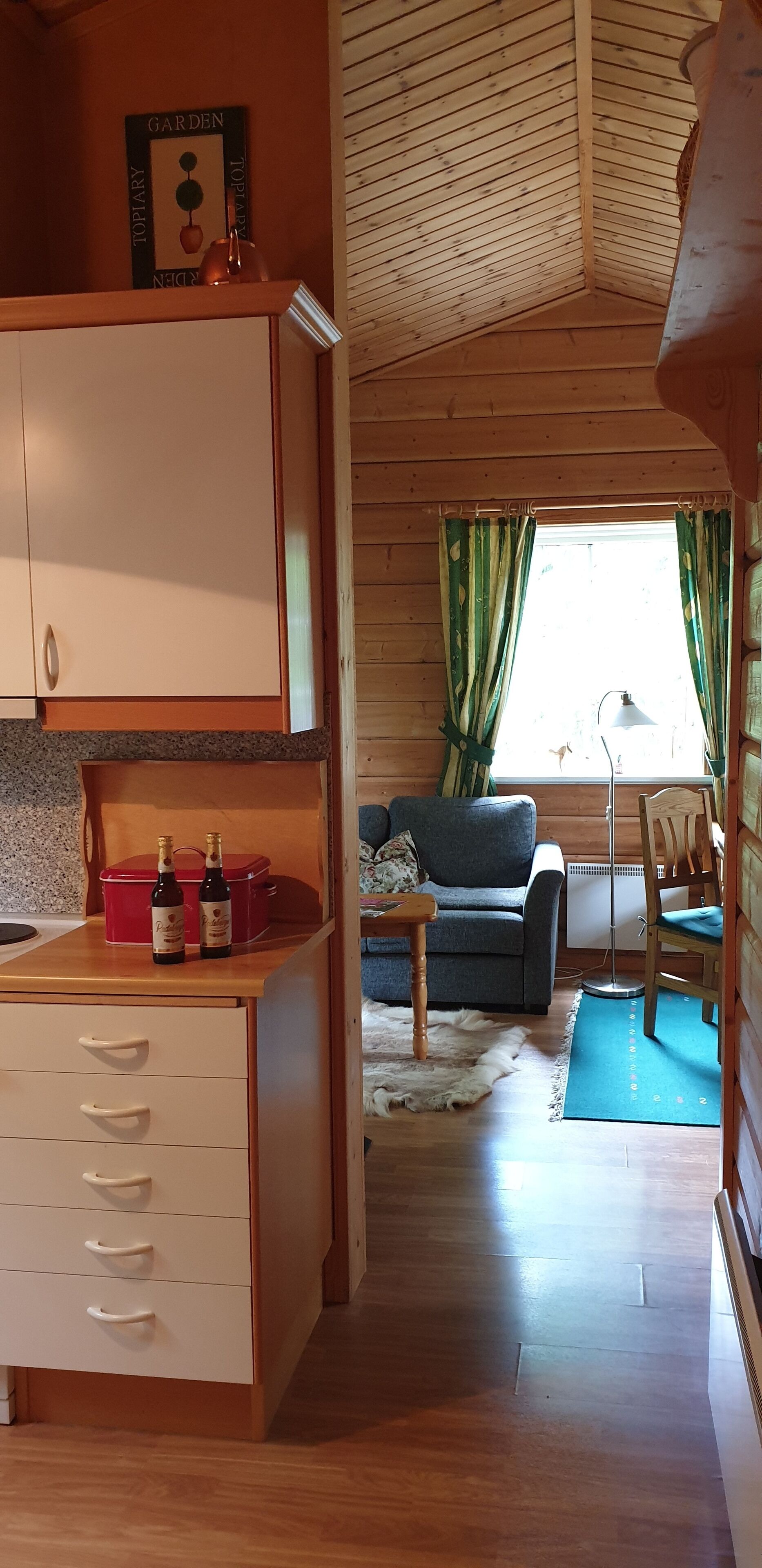 L’intérieur chaleureux d’un chalet avec des murs en bois, un coin cuisine doté de placards, une table à manger avec des bouteilles, un canapé et une fenêtre avec des rideaux.