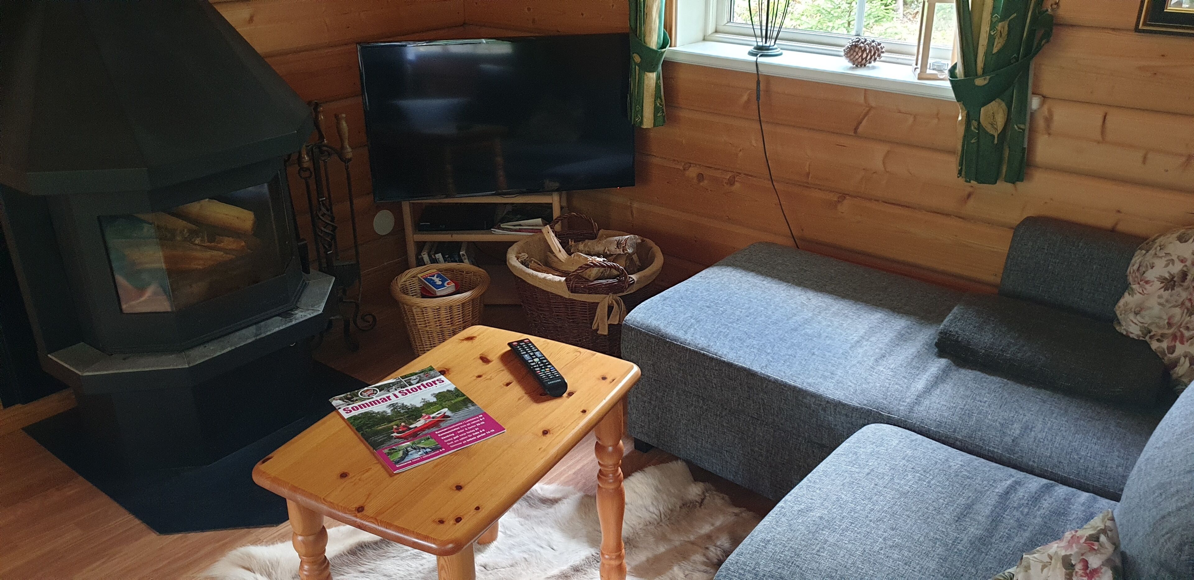 L’intérieur d’un chalet accueillant, avec un poêle à bois, un téléviseur posé sur un meuble, un canapé gris et une petite table sur laquelle se trouvent un livre et une télécommande.