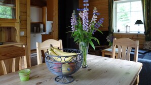 Ein Interieur einer Holzhütte mit Esstisch, Stühlen, einer Obstschale, einer Blumenvase und einem Fenster mit Vorhängen.