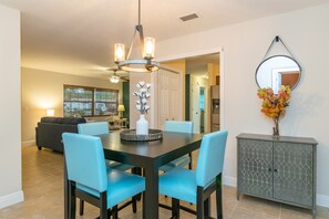 A dining area with a dark wooden table, light blue chairs, and a chandelier. There is a cabinet with a decorative mirror and a vase with flowers.