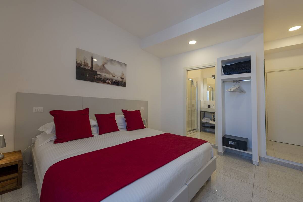 A modern hotel room with a large bed, red pillows, a painting on the wall, a walk-in closet, and a bedside table with a lamp.