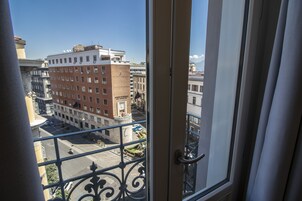 Une vue depuis un balcon doté d’une rambarde en métal, donnant sur une rue de la ville avec des immeubles et des voitures garées.