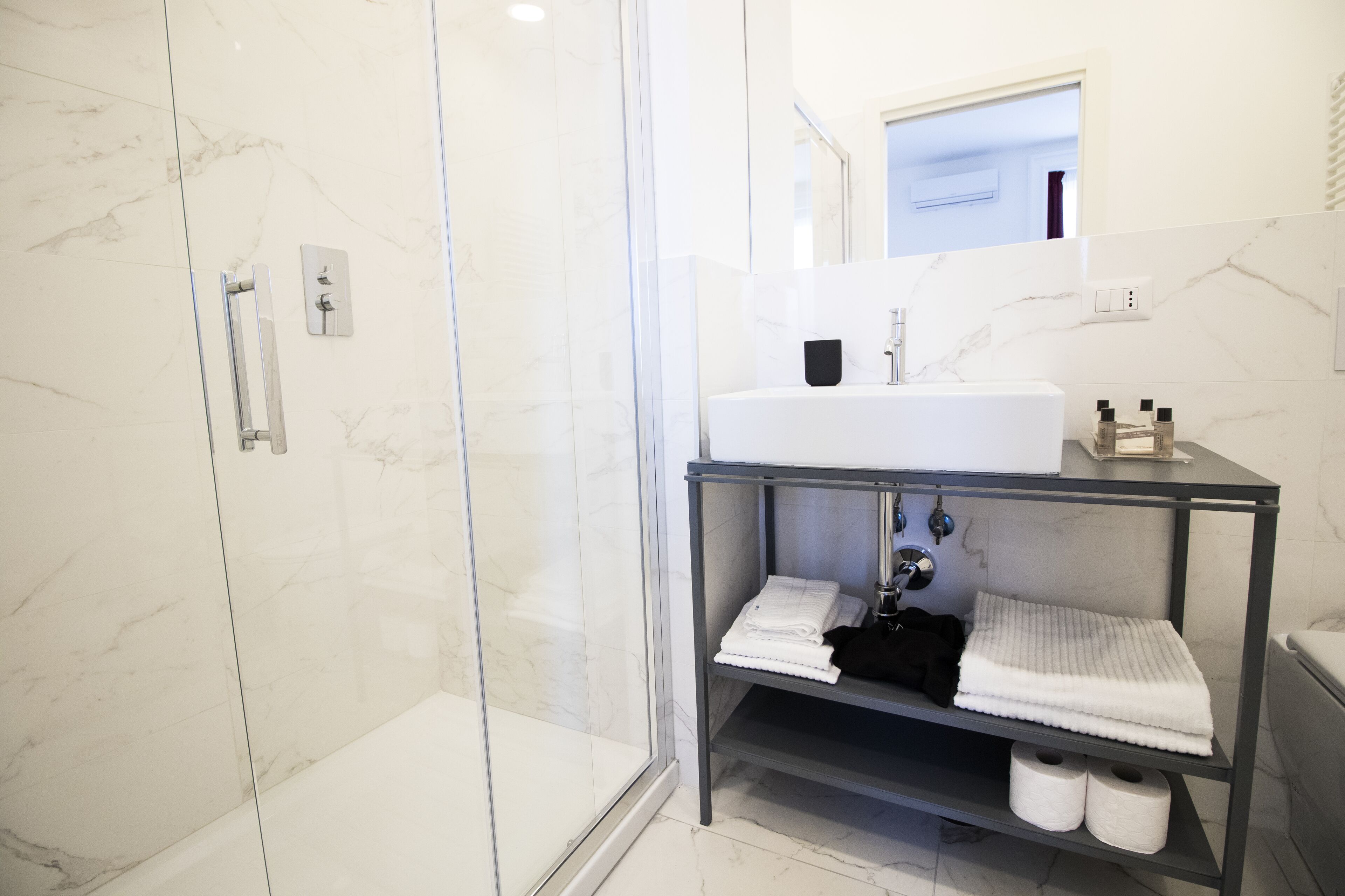 A modern bathroom with a glass shower enclosure, a white sink with a marble countertop, and a shelf with towels and toiletries.