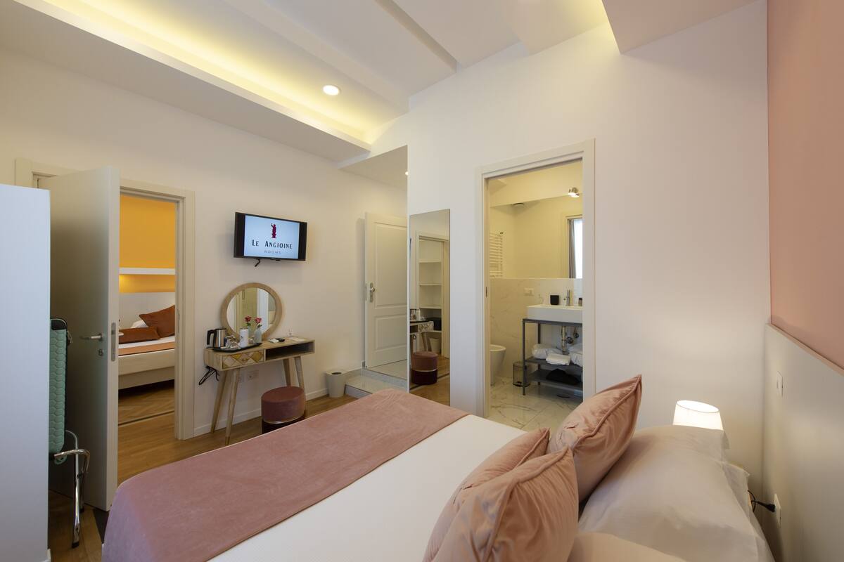 A modern hotel room with a bed, a vanity with a mirror, a television, and a bathroom visible through an open door.