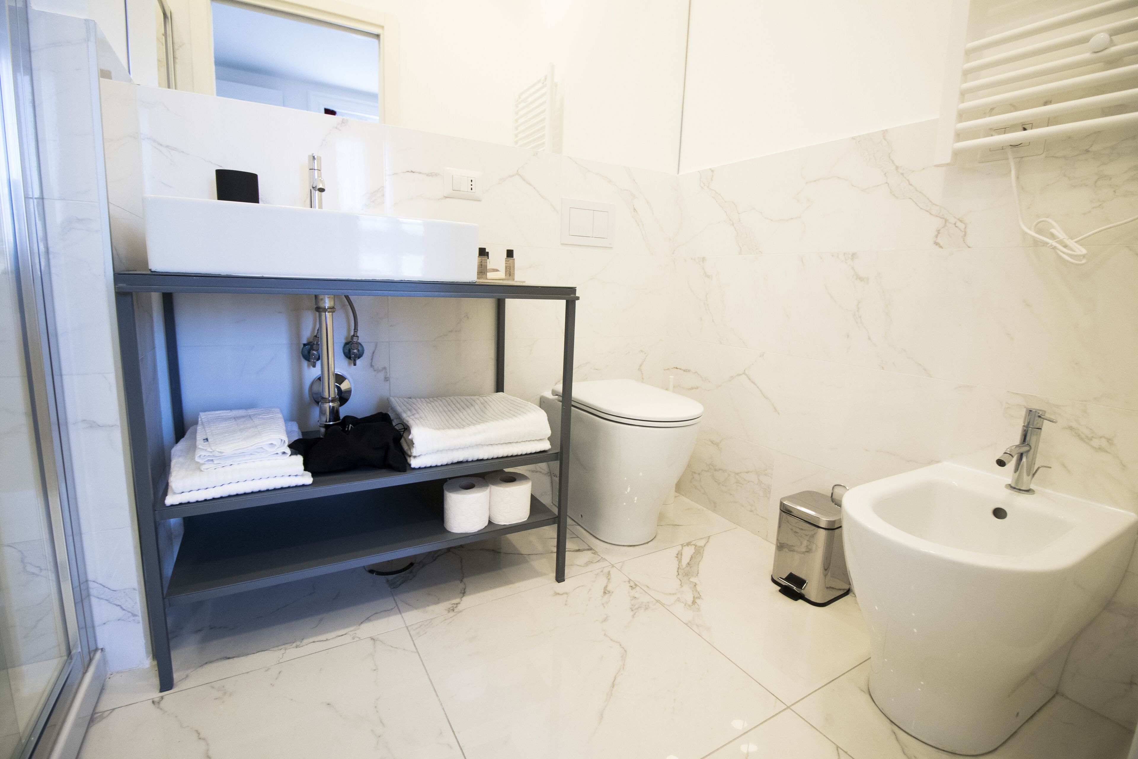 A modern bathroom with a marble-tiled floor, a white sink, a bidet, and a shelf with towels and toiletries.
