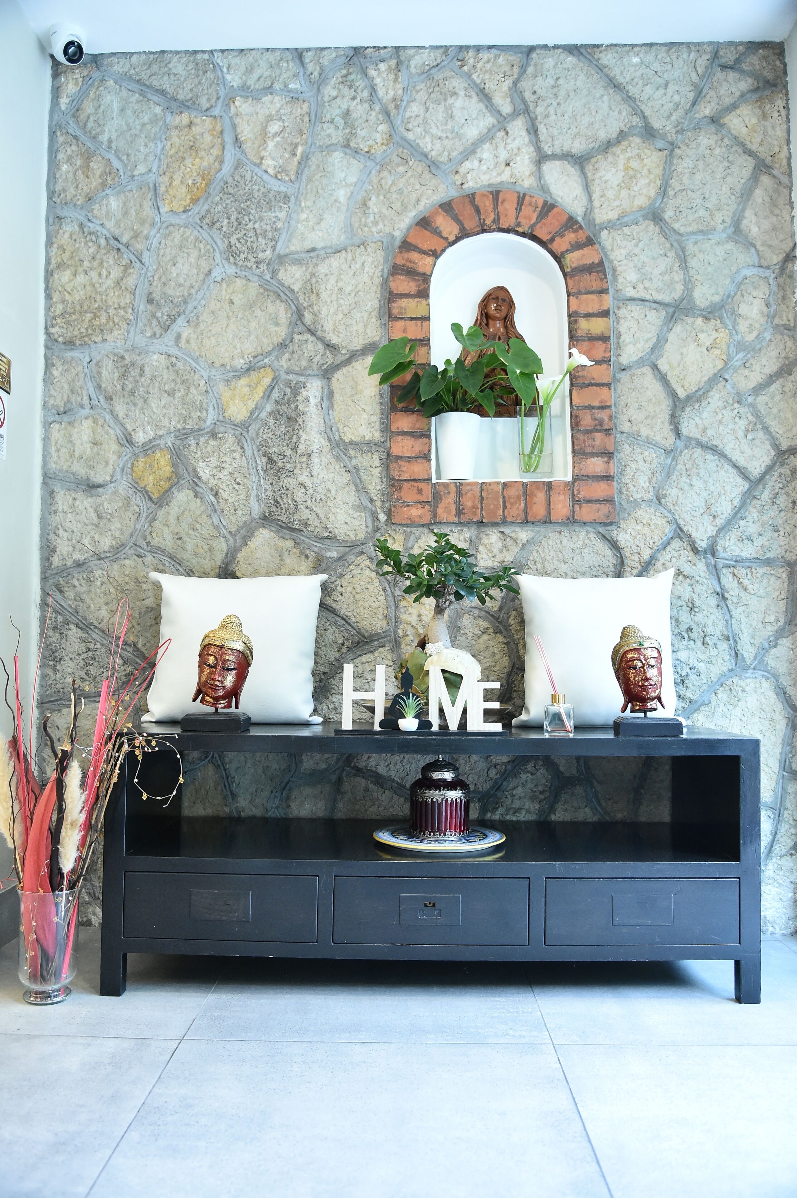 Una consola negra con pared de piedra, dos cojines blancos con estatuas de Buda, un espejo y la palabra “HOME” sobre la mesa.