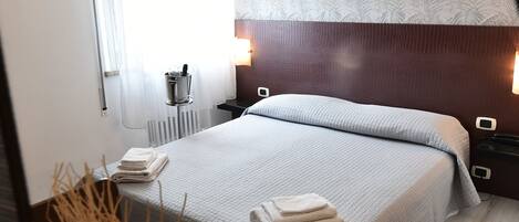 A neatly made bed with a white comforter and pillows, a bedside table with a lamp, and a radiator on the wall.