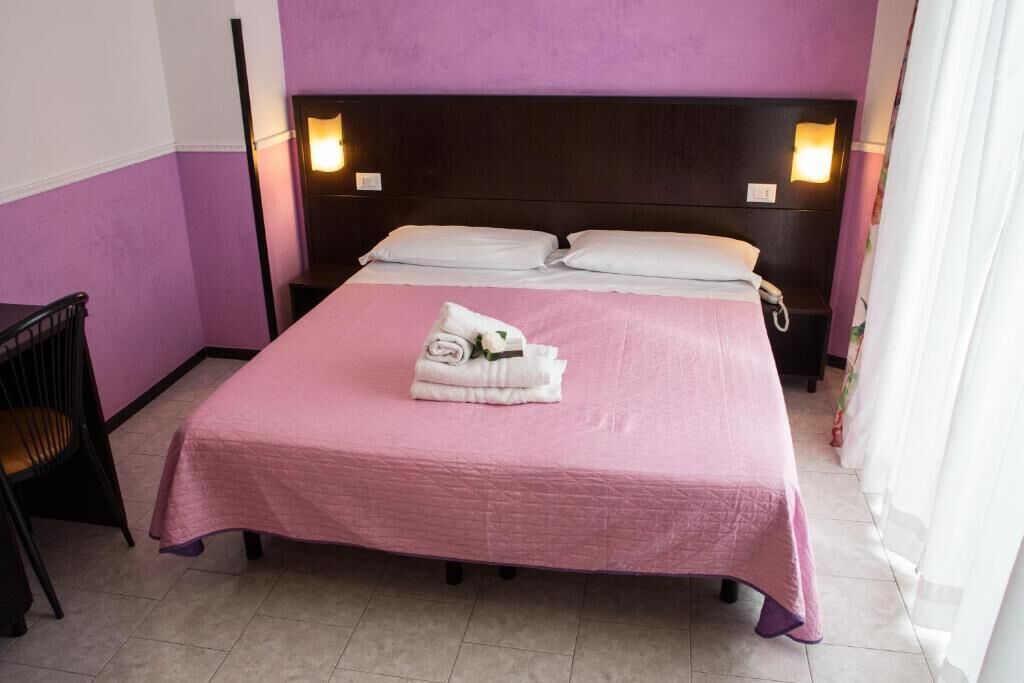 A neatly made bed with a pink bedspread and white pillows, a bedside table with a lamp, and a chair in a room with purple walls.
