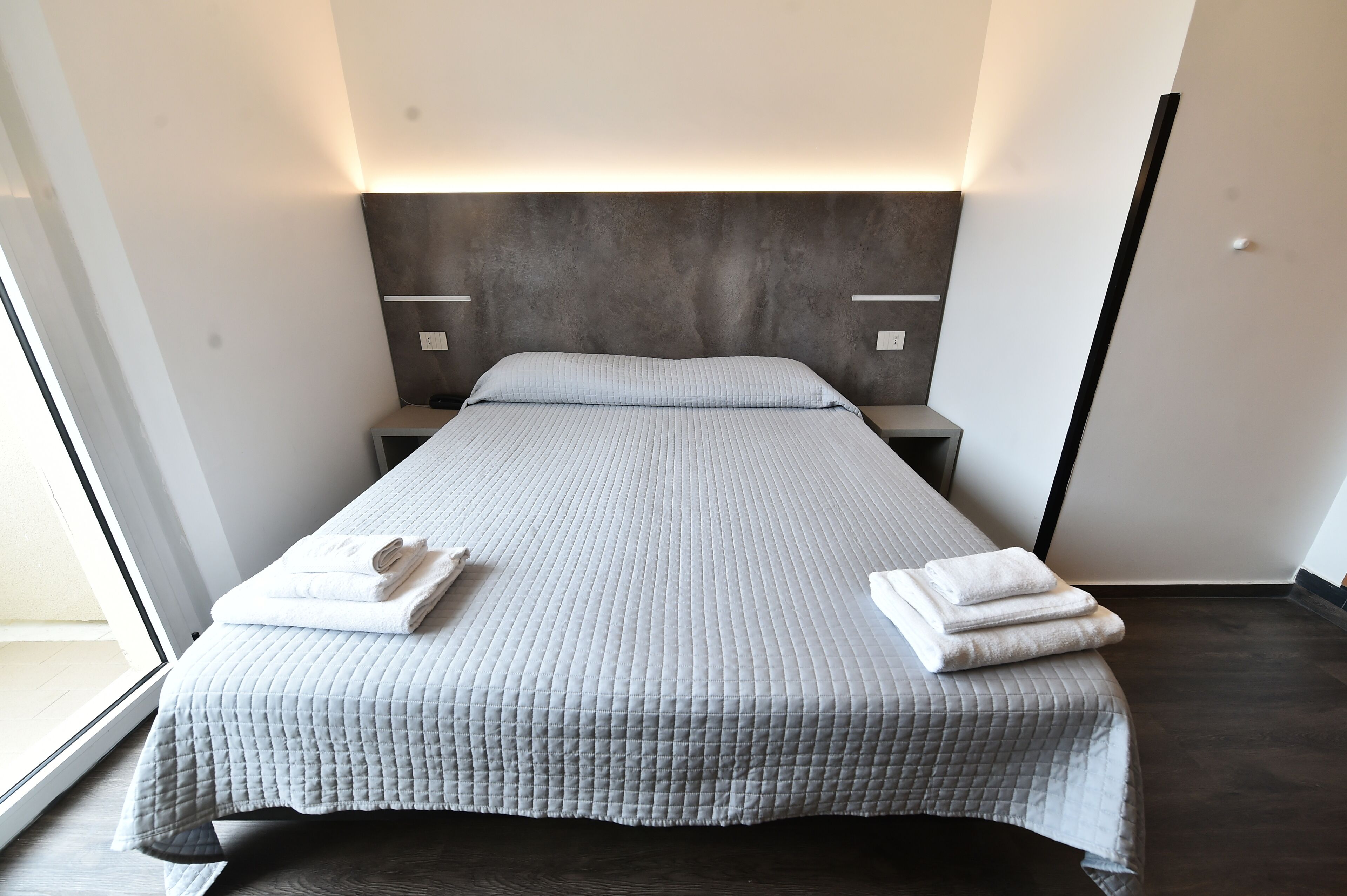 A neatly made bed with a grey quilt, two white towels, and a modern headboard.