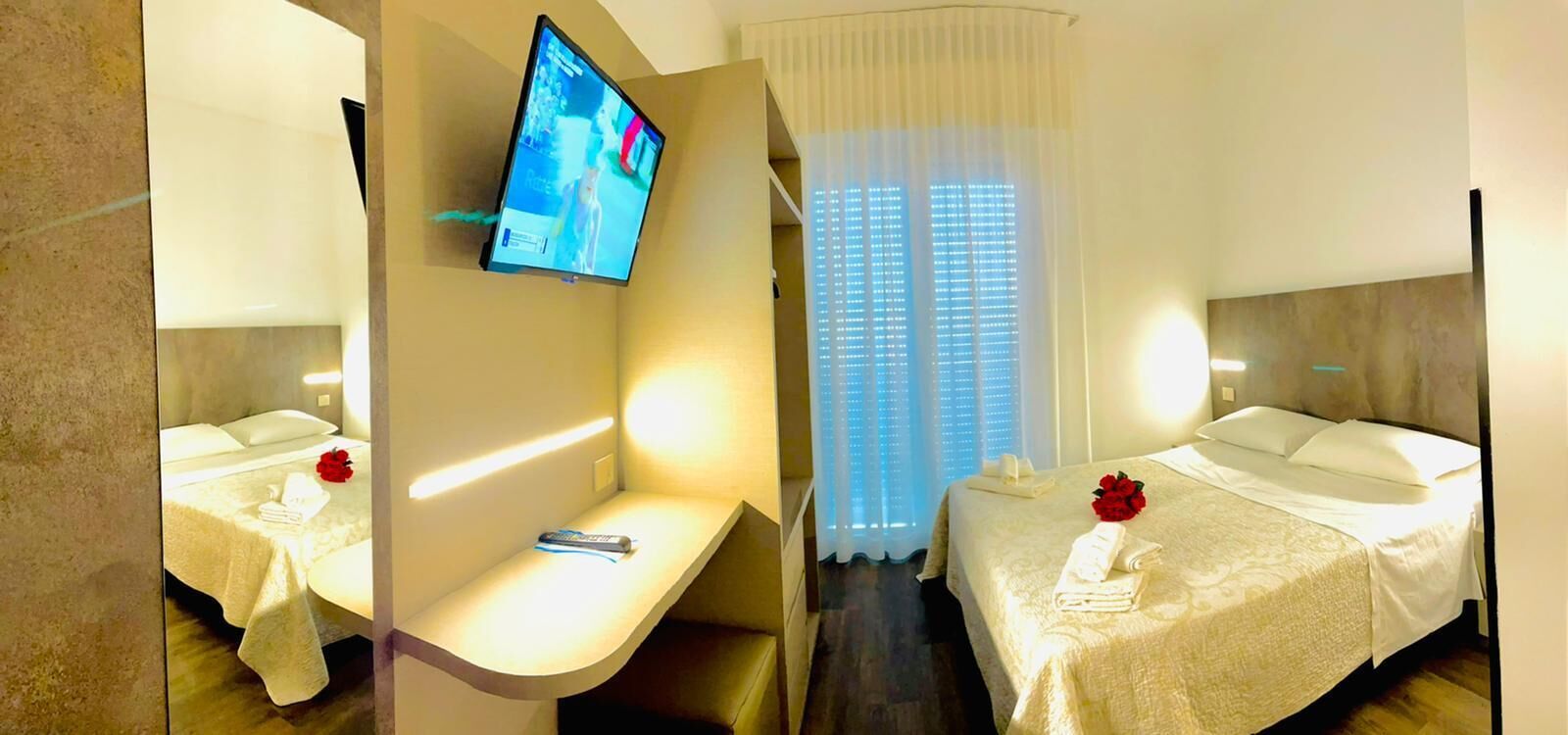 Une chambre d’hôtel avec un lit, un téléviseur fixé au mur, une table de chevet et une fenêtre avec des rideaux.