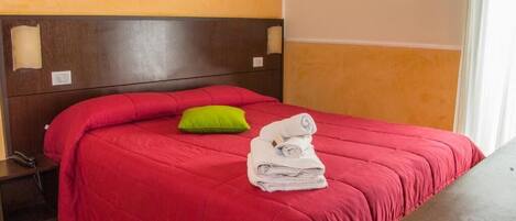 A hotel room with a red bedspread, a green pillow, and a stack of white towels on the bed.