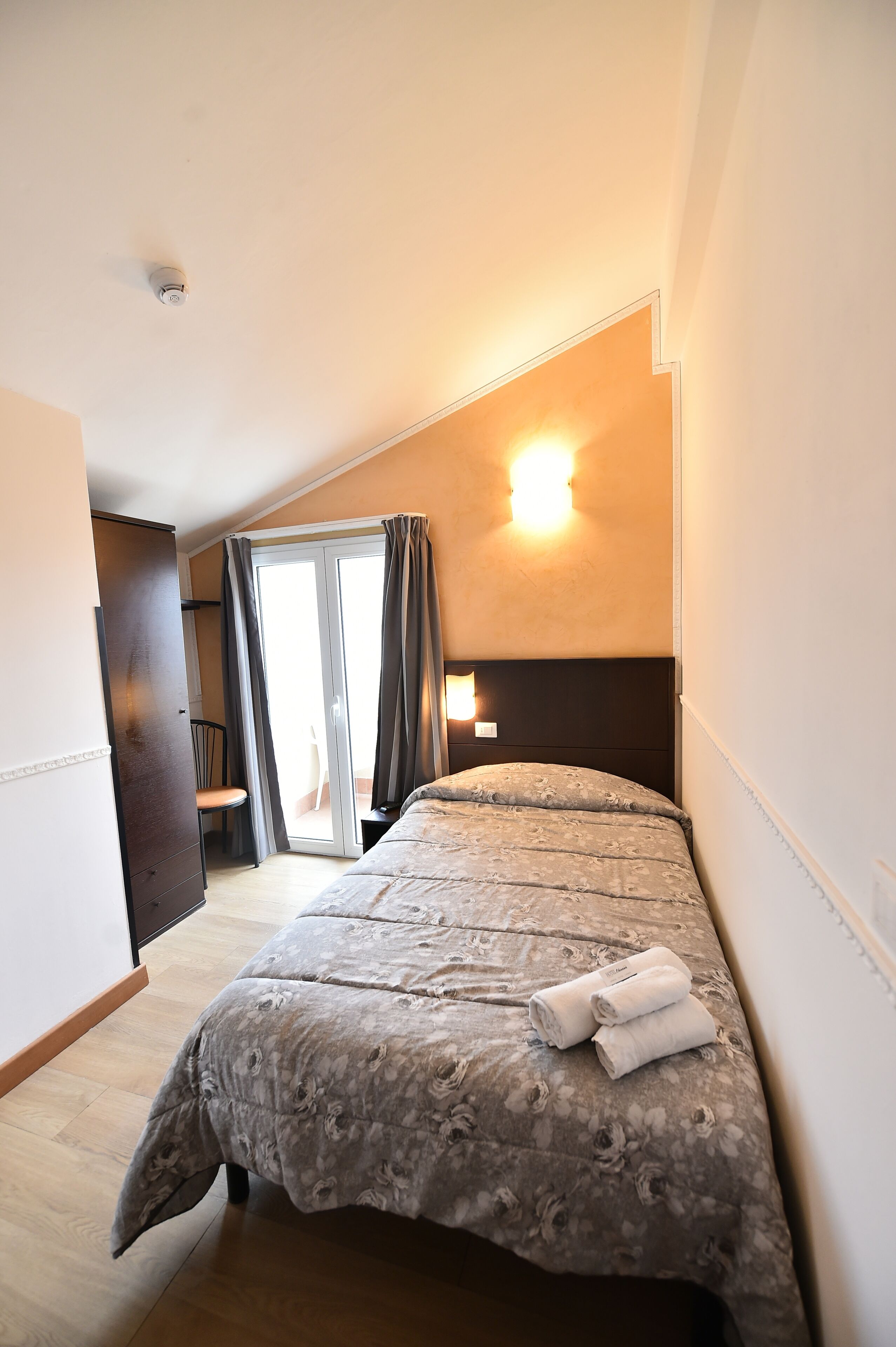 Una camera da letto con un letto, un comodino, una sedia, un armadio e una porta vetrata scorrevole che dà su un balcone.