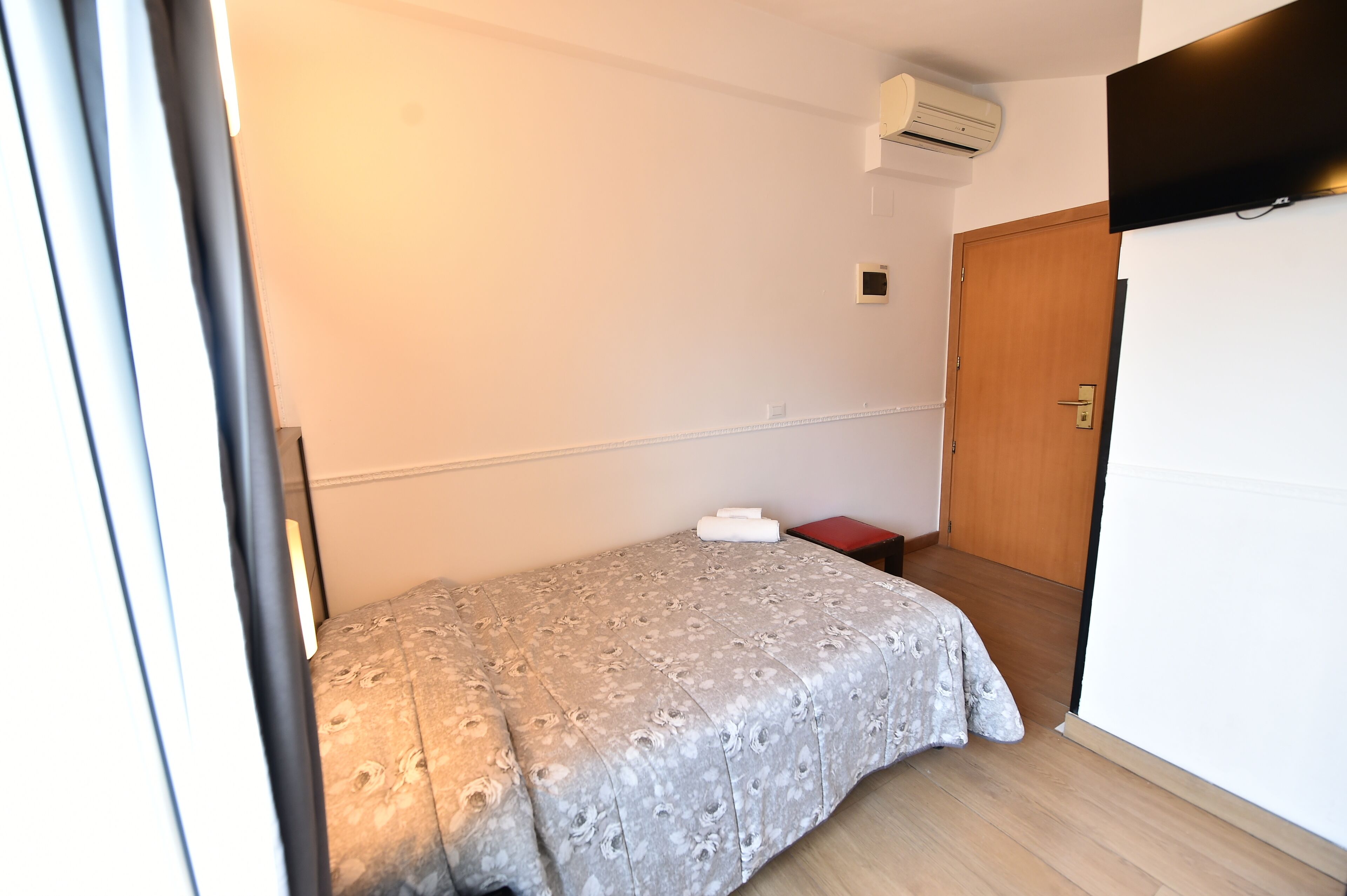 A small, neatly arranged bedroom with a bed, a wooden door, and a wall-mounted air conditioner.