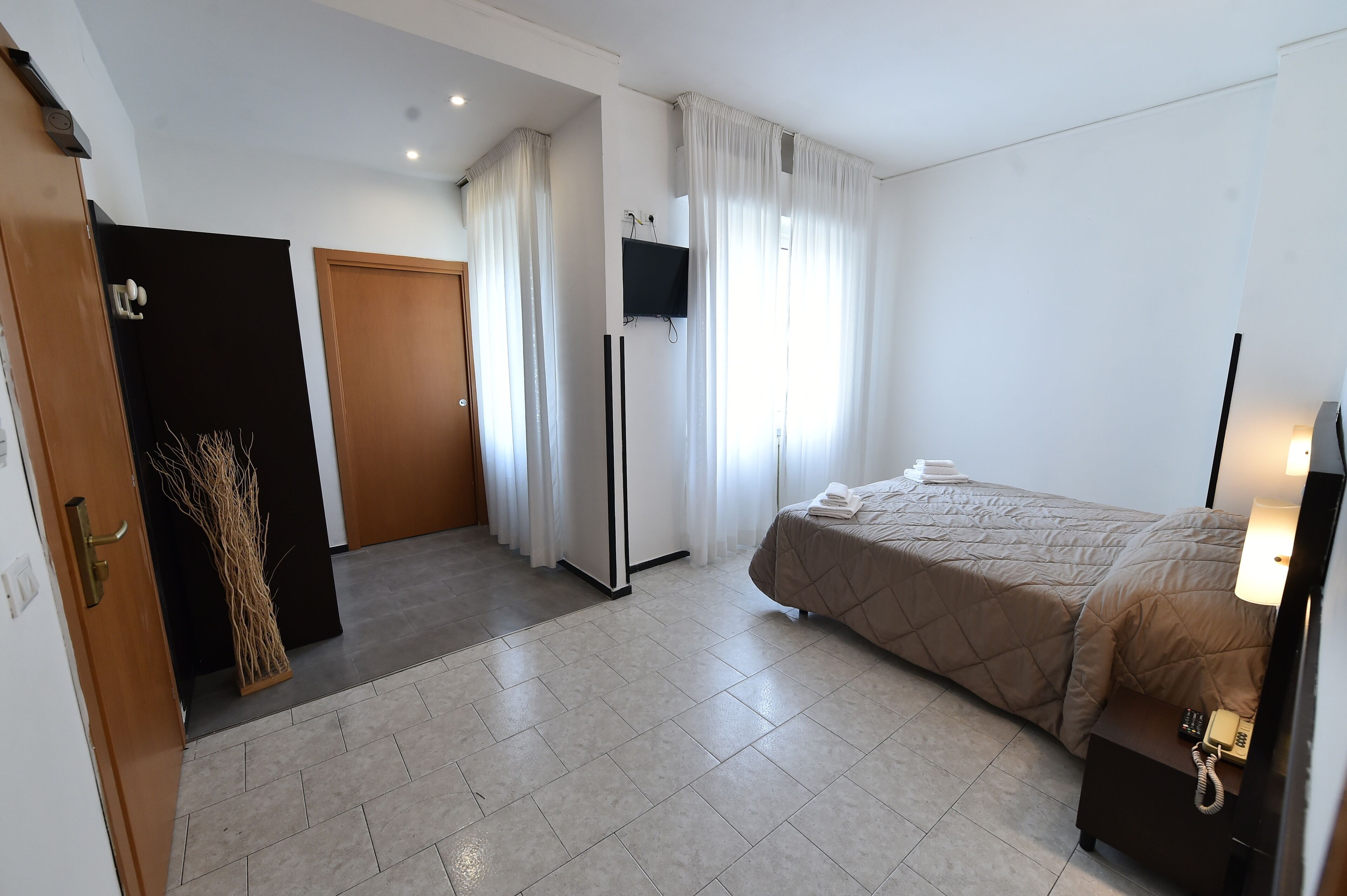 A hotel room with a bed, a desk with a lamp, a TV mounted on the wall, and a door with a towel on it.