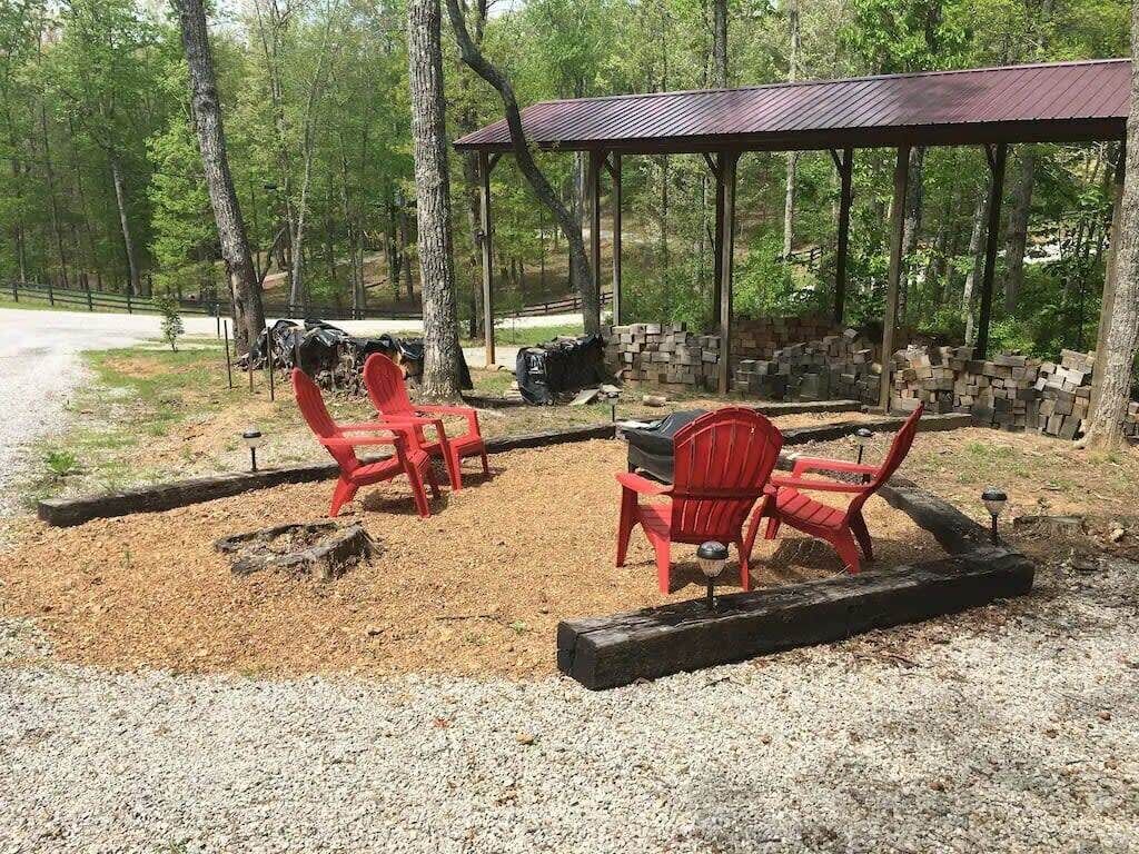 A covered outdoor seating area with red chairs and a grill, surrounded by trees and gravel.