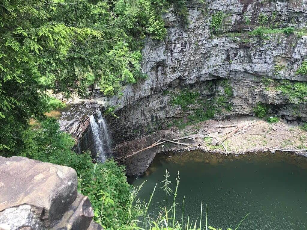 Een natuurlijke waterval die in een kalm stroompje uitmondt, omringd door rotswanden en weelderige begroeiing.