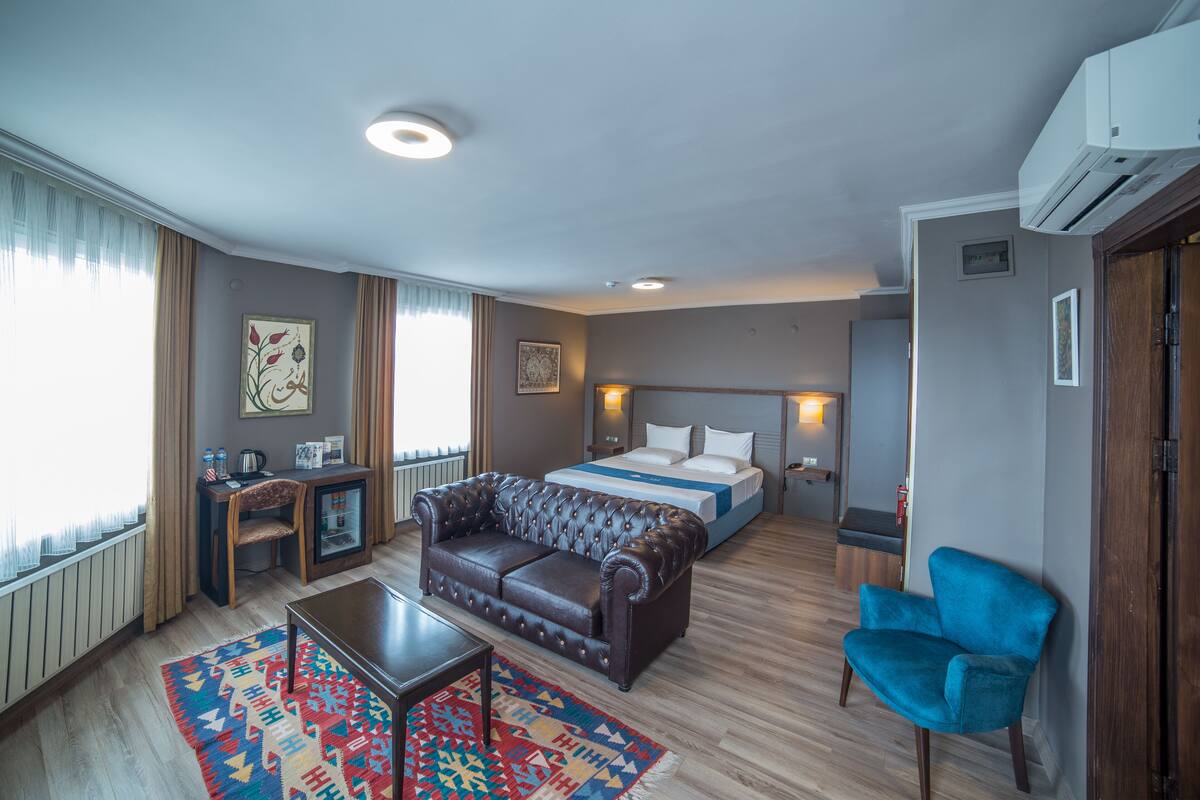 Una camera d'albergo con un letto, un divano in pelle, una poltrona blu, un tavolino e un tappeto colorato.