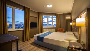 Habitación de hotel con una cama grande, un escritorio, una silla, un televisor y vistas a la ciudad desde la ventana.