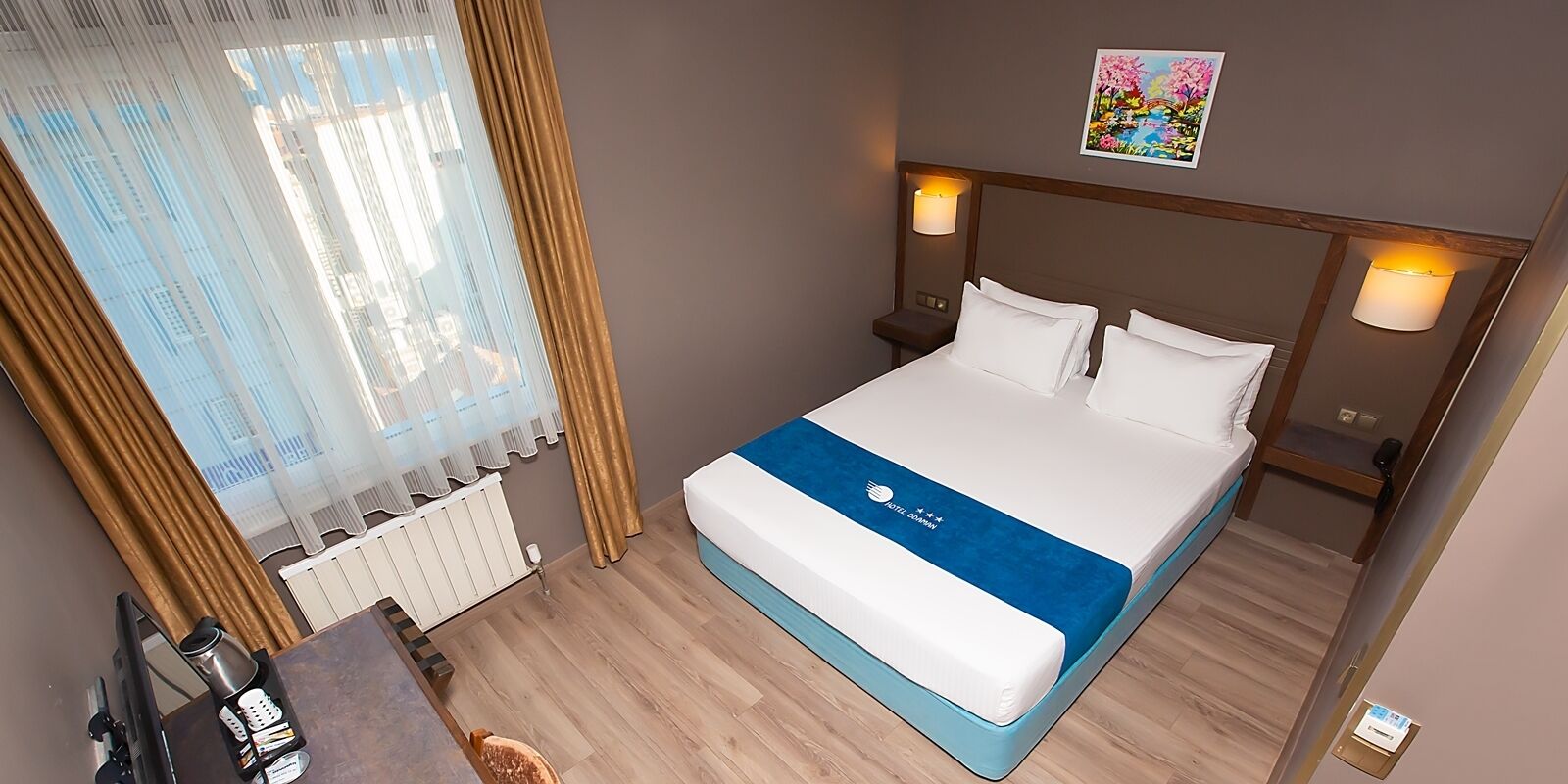 Habitación de hotel con cama, lámparas de noche, radiador, ventana con cortinas y un cuadro en la pared.