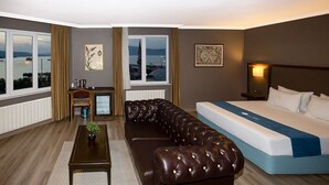 Habitación de hotel con cama, sofá de cuero, una mesita con una planta y vista al mar a través de amplios ventanales.