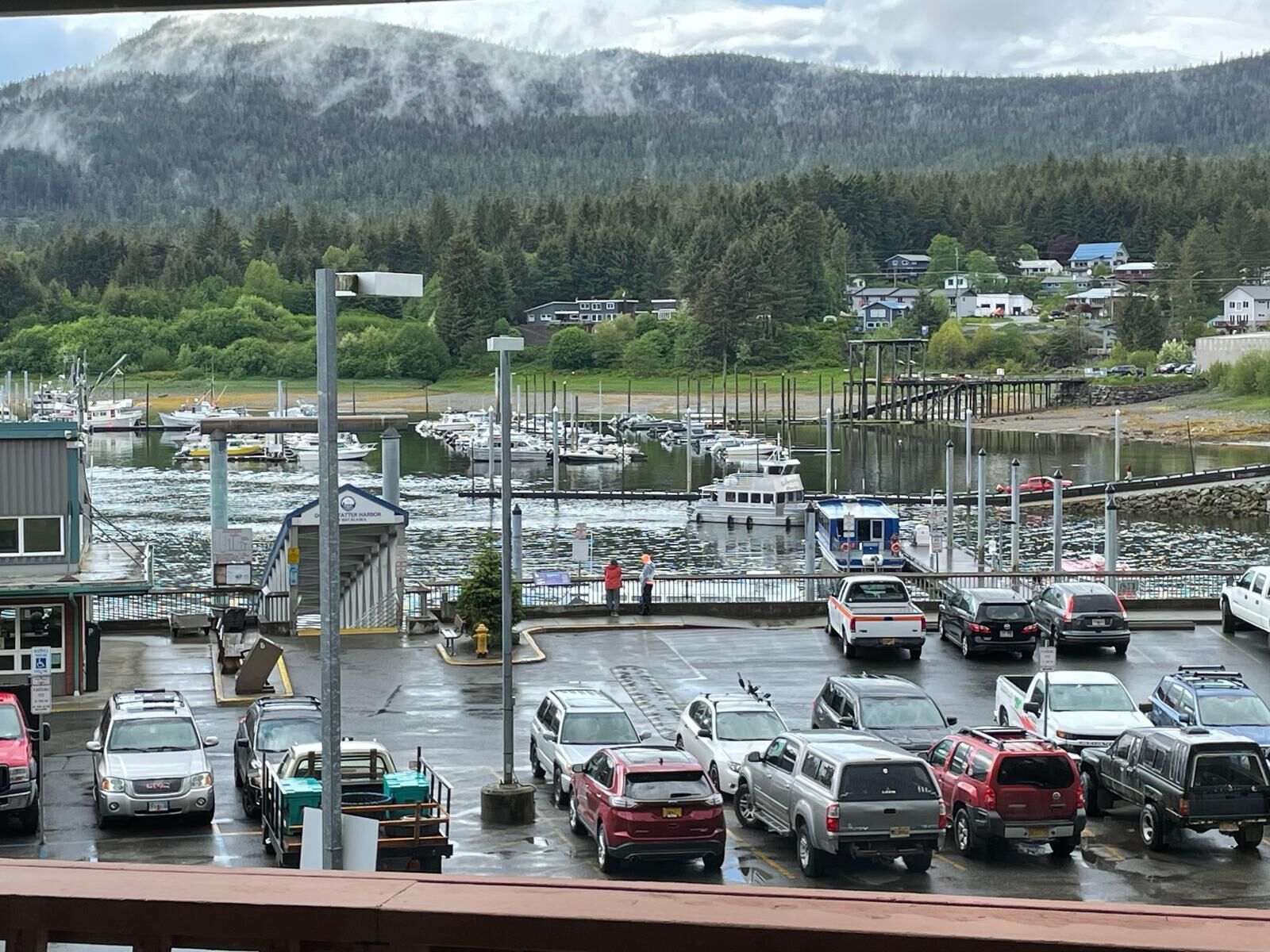 A marina with boats docked, a parking area with cars, and a mountainous backdrop.