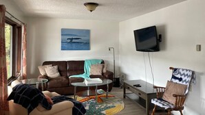 Uma sala de estar com um sofá marrom, uma mesa de centro de vidro, uma cadeira de madeira, uma TV fixada na parede e uma janela com cortinas.