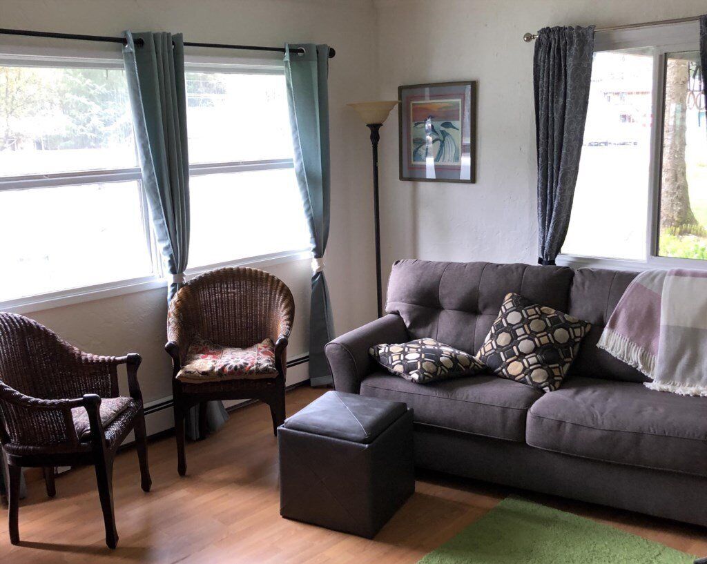 Ein Wohnzimmer mit einem grauen Sofa, zwei Korbstühlen, einem schwarzen Hocker und einem grünen Teppich.