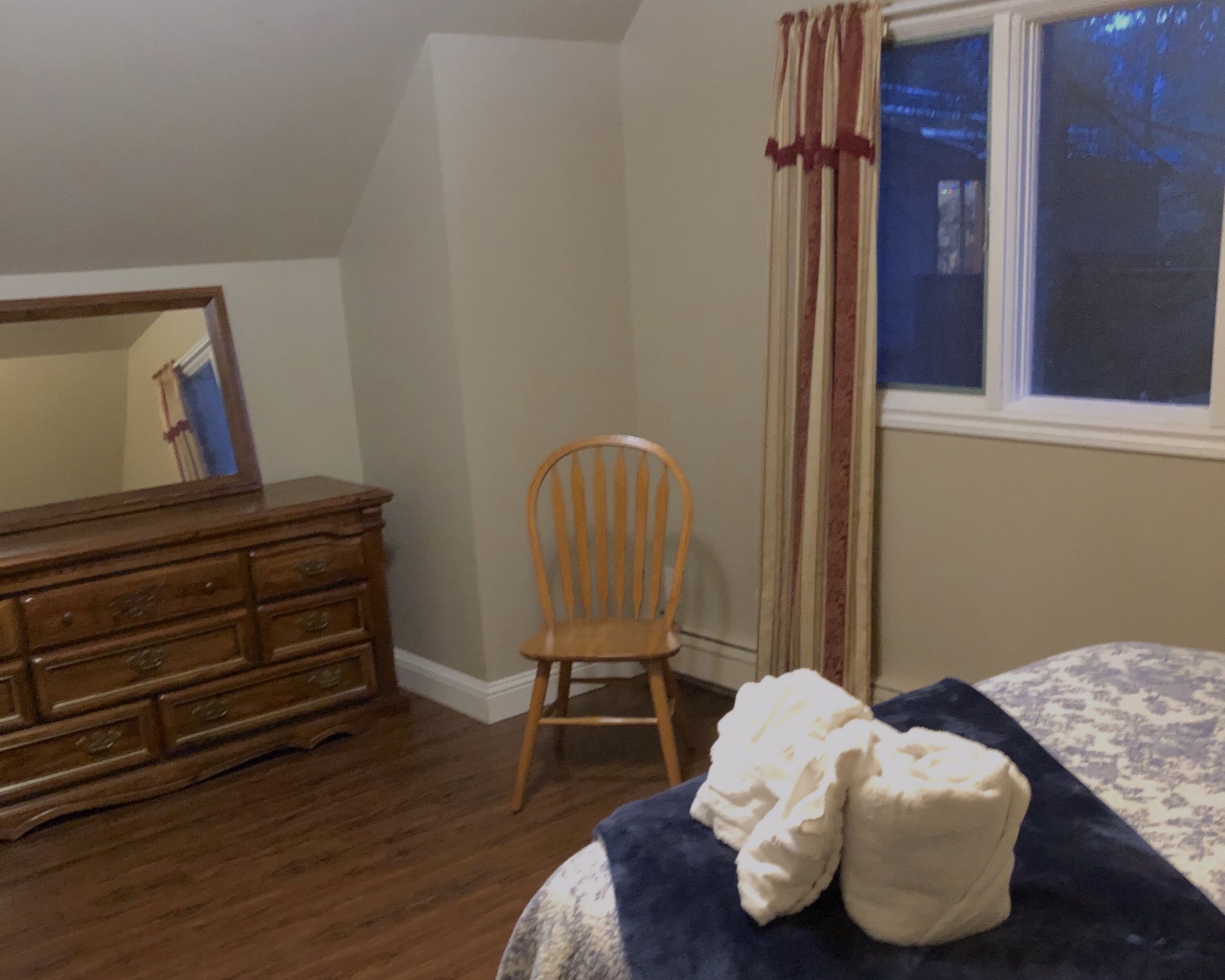 A bedroom with a wooden dresser, a chair, a bed with a blue blanket, and a window with curtains.
