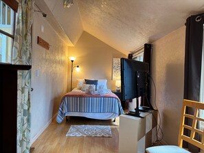 Ein Schlafzimmer mit Bett, Fernseher, Schreibtisch, Stuhl, Lampe und einem Fenster mit Vorhängen.